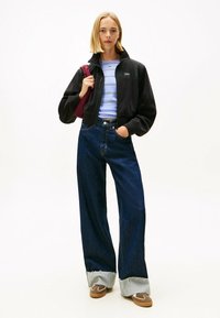 Giacca in pile nera con zip, camicia a righe azzurro chiaro, jeans scuri a gamba larga con orli sfrangiati e sneaker marroni. Borsa a tracolla bordeaux.