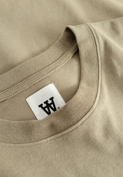 T-shirt en coton beige avec un col rond, présentant une texture douce et une étiquette avec une écriture noire placée près de la couture du col.