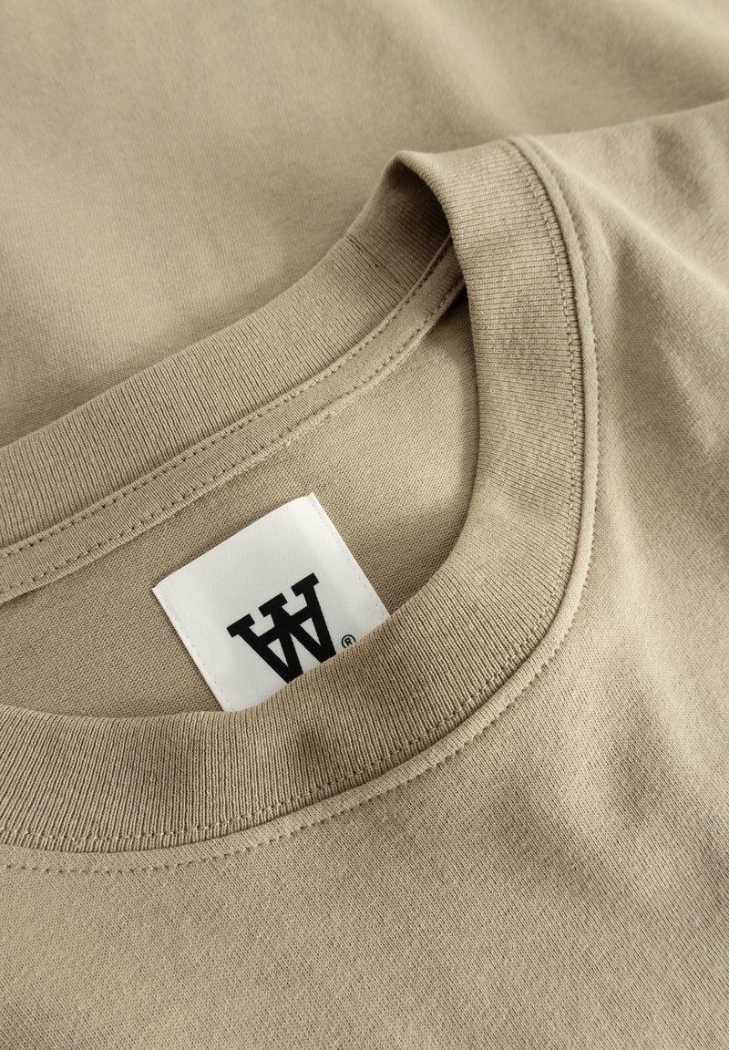 Camiseta de algodón beige con escote redondo, que presenta una textura suave y una etiqueta con letras negras situada cerca de la costura del cuello.