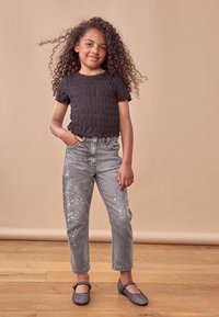 Top corto a maniche corte nera con smock, jeans grigi con dettagli di paillettes e ballerine grigie scintillanti. Sullo sfondo c'è una tonalità neutra.