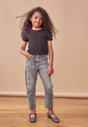 Svart smockad kortärmad topp, grå jeans med paljetter, och grå glittriga ballerinaskor. Bakgrunden har en neutral ton.