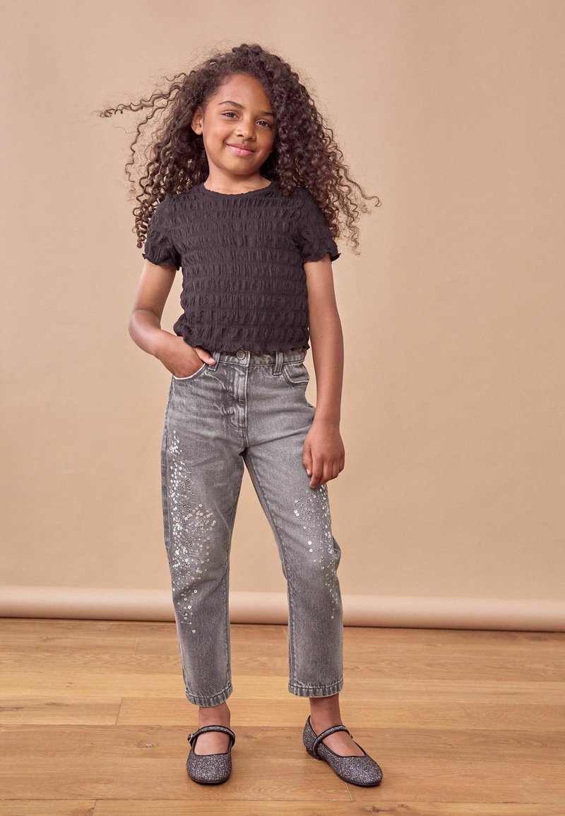 Top corto a maniche corte nera con smock, jeans grigi con dettagli di paillettes e ballerine grigie scintillanti. Sullo sfondo c'è una tonalità neutra.