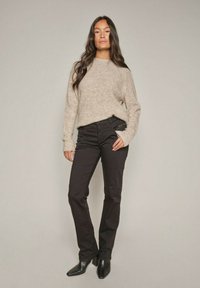 Helles beige Strickpullover mit gerippten Akzenten, kombiniert mit schmalen schwarzen Hosen und schwarzen spitzen Knöchelstiefeln, vor einem neutralen Hintergrund.