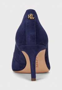 Tummansininen mokkanahkainen korkokenkä, jossa on terävät kärjet, kultainen logovivahde kantapäässä ja ohut stiletto, joka nostaa suunnittelua.