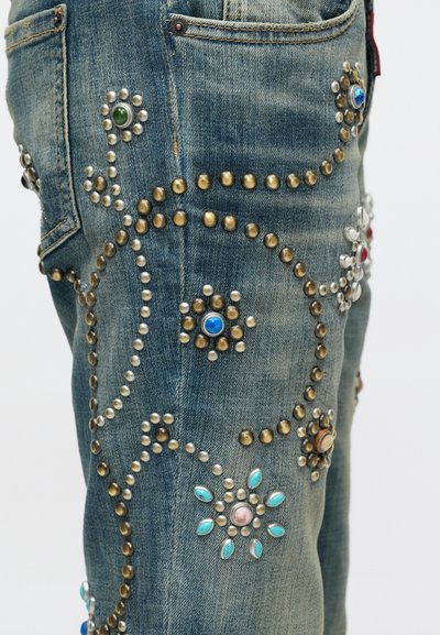 Džínsy z denimu s kvetinovými a kruhovými kovovými nitmi a farebnými kameňmi usporiadanými v dekoratívnych vzoroch na stehnách a v oblastiach vreciek.