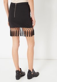 Minifalda negra de diseño entallado, con cremallera trasera y detalle de red con flecos en el dobladillo. Acentuada con borlas negras colgantes.