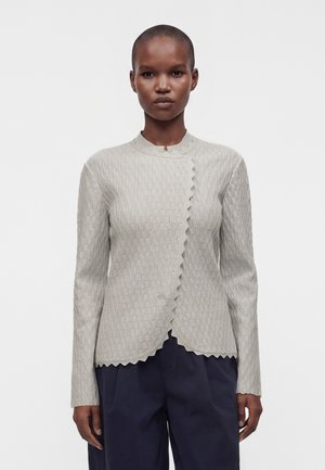 Persoon staat en kijkt vooruit, draagt een lichtgrijze, getextureerde lange mouwen top met geschulpte randen en een donkere broek tegen een effen achtergrond.