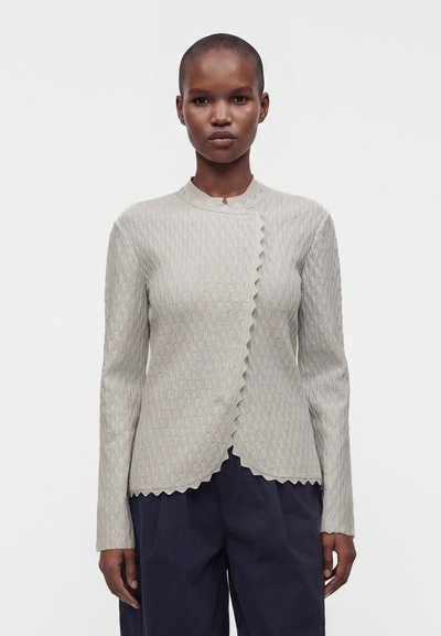 Personne debout, de face, portant un haut à manches longues gris clair texturé avec des bords festonnés et un pantalon foncé sur un fond uni.