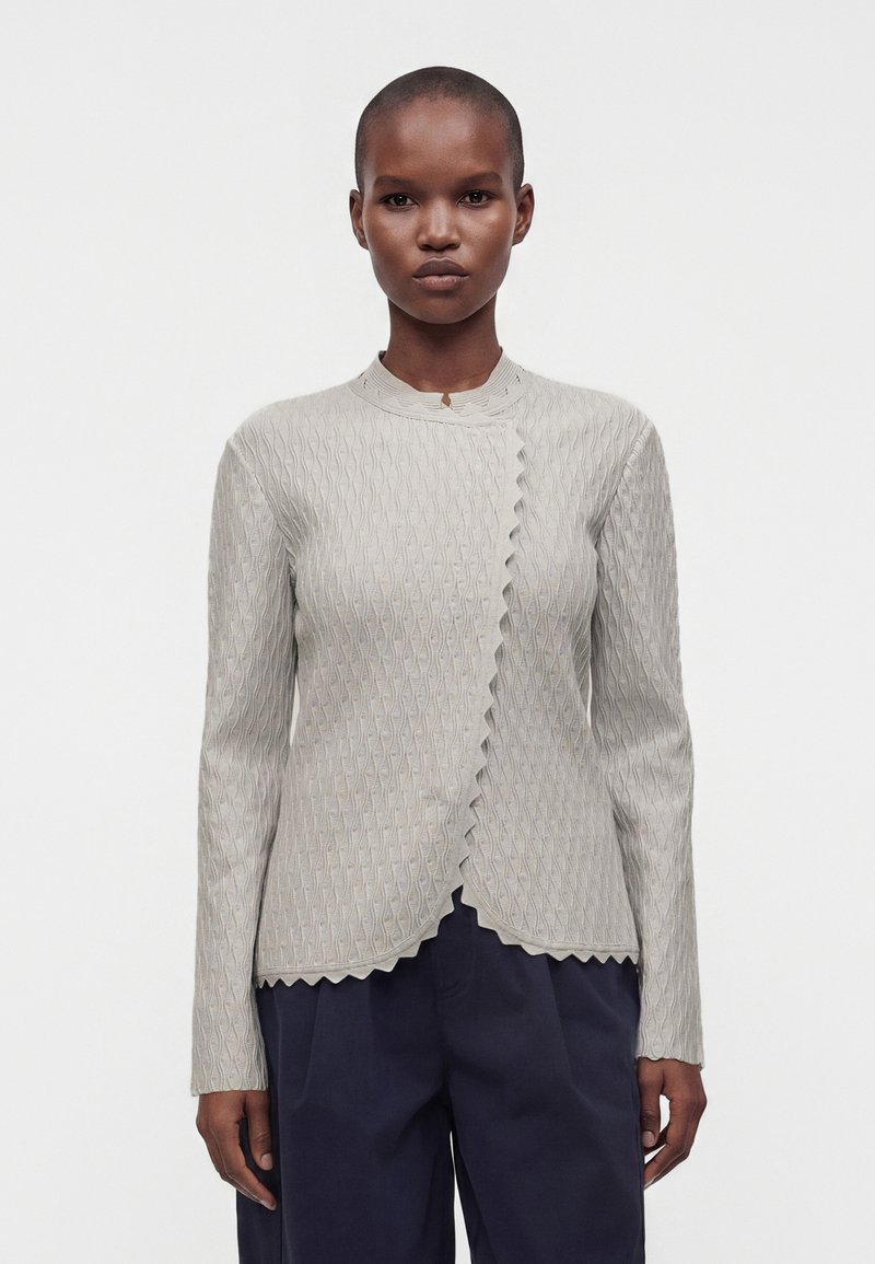 Personne debout, de face, portant un haut à manches longues gris clair texturé avec des bords festonnés et un pantalon foncé sur un fond uni.