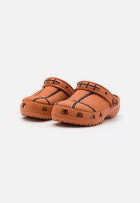 Saboti portocalii din cauciuc, cu o suprafață texturată, accente negre și găuri de ventilație. Prezintă o curea ajustabilă și un design cu toc înalt.