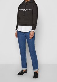 Cappuccio nero corto con logo bianco, indossato sopra una camicia bianca con bottoni. Jeans in denim blu con orli arrotolati e mocassini neri.
