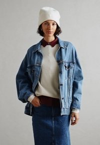 Persona, kas uzvilkusi baltu beanie, slāņotu džinsu jaku, krēmkrāsas svīteri, bordo krekls un džinsu svārkus, stāv ar vienu roku kabatā pret vienkāršu fona.