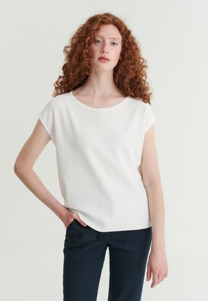 Frau mit lockigem roten Haar, die ein weißes kurzärmliges Oberteil und dunkle Hose trägt, steht mit einer Hand in der Tasche vor einem schlichten Hintergrund.