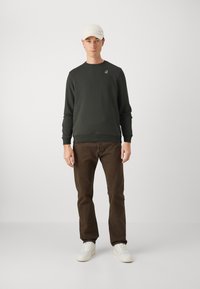 Felpa verde scuro con polsini e orlo a costine, logo ricamato. Pantaloni marroni e sneakers bianche. Un cappellino bianco completa l'outfit.