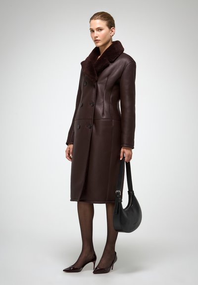 Abrigo de cuero marrón con un cuello de piel de contraste, diseño de doble botonadura y silueta elegante. La mujer sostiene un bolso negro y lleva zapatos de tacón.