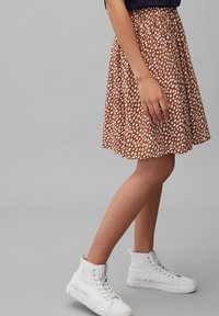 Gonna marrone con macchie bianche, elastico in vita, design svasato. Abbinata a sneakers bianche alte. Stile minimalista e casual.
