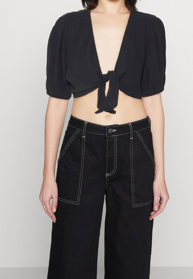 Blouse noire courte avec des manches ballon et un lien à l'avant, associée à un pantalon noir à jambes larges avec des coutures blanches et des poches avant.