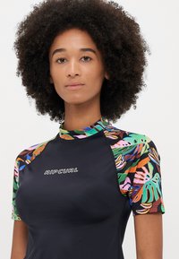 Kombinezon z krótkim rękawem, czarnym korpusem i kolorowym tropikalnym wzorem liści na rękawach i kołnierzu; na klatce piersiowej znajduje się logo "RIPCURL".