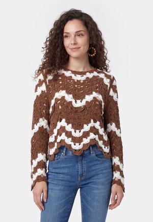 Kvinde med krøllet brunt hår iført brun og hvid hækletrøje og blå jeans, stående mod en ensfarvet lys baggrund.