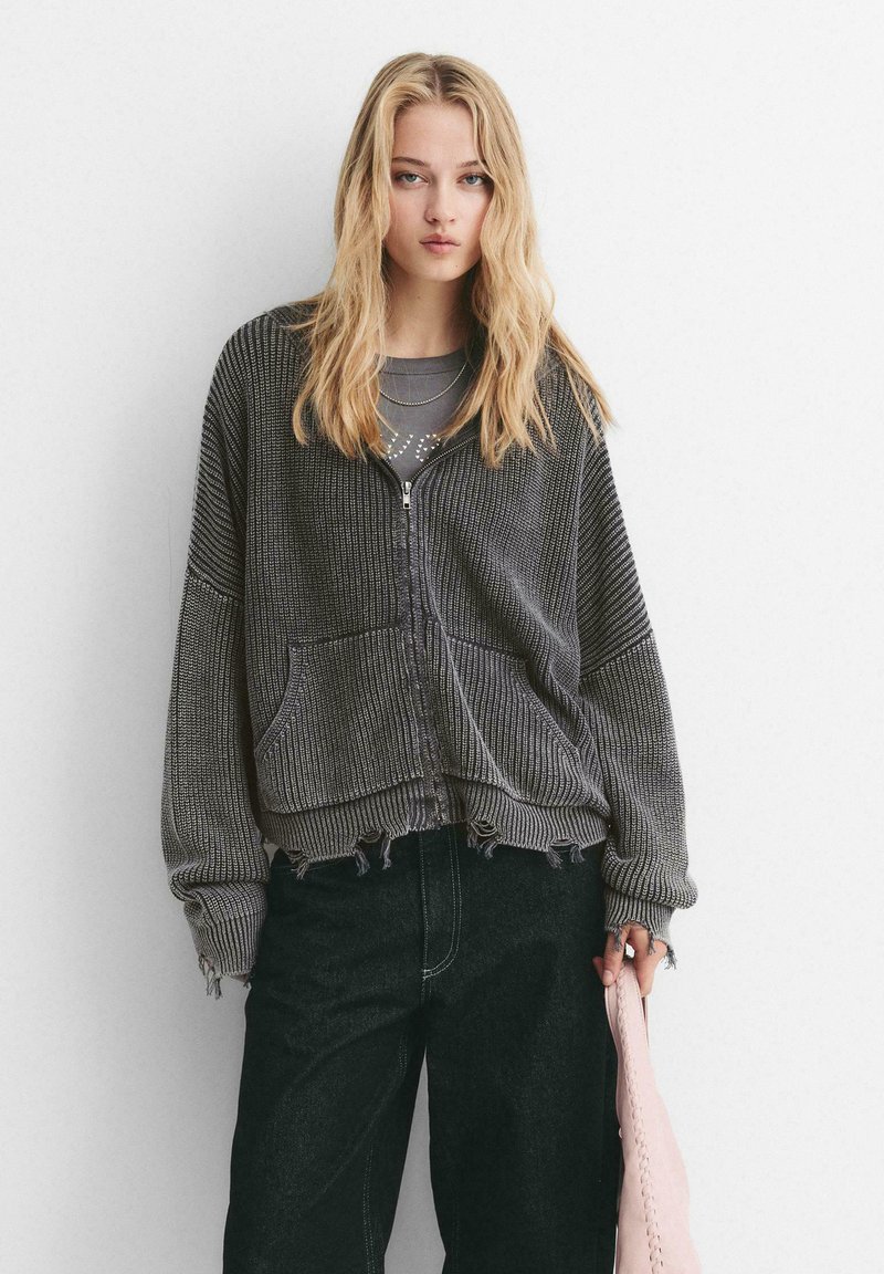 Grå strikket zip-up cardigan med et cropped design, der har en tekstureret overflade, frontlommer og rå kant. Sat sammen med mørke jeans og en pink taske.