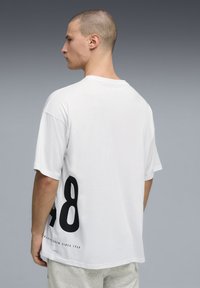 T-shirt en coton blanc avec un grand numéro 48 noir sur le côté gauche et un texte près de l'ourlet, coupe décontractée avec manches courtes et col rond.