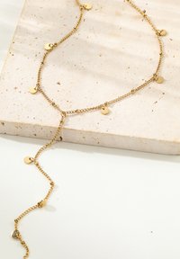 Collier doré avec chaîne maillée et pendentifs ronds, doté d'un long cordon ajustable. Sur fond texturé de couleur neutre.