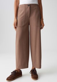 Bruine wijde broek met een gladde textuur, voorzien van een knoopsluiting aan de voorkant, twee voorzakken en een getailleerde snit. Draagt met bruine sneakers.