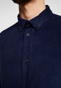 Laivastonsininen corduroy-paita, jossa on napillinen kaulus, mustat napit ja teksturoitu kangas, korostaen sileää pintaa ja hienoa ribbikuviota.