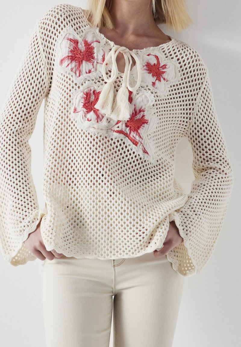 Femme portant un pull en crochet crème avec une broderie florale rouge et des liens à pompons, associé à un pantalon ajusté de couleur claire.