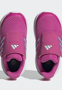 Zapatillas deportivas moradas con un upper de tela texturizada, correa ajustable y acentos blancos. La suela es redondeada con un diseño ligero.