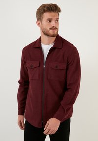 Burgundy színű, zipzáros dzseki struktúrált anyagból, gallérral, két gombos elülső zsebbel és testhez álló szabással.