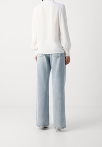 Person iført en hvid langærmet bluse og lyseblå brede jeans, stående mod en ensfarvet hvid baggrund.