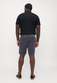 Herre svart polo skjorte med kortermede ermer, kombinert med grå shorts. Shortsen har en glatt tekstur og to baklommer, iført svarte sko.