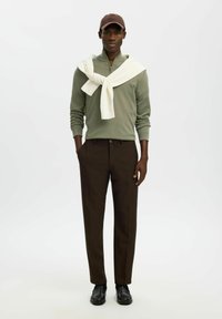 Homme portant une casquette marron, un pull à fermeture éclair vert avec un pull blanc drapé sur les épaules, un pantalon marron foncé et des chaussures noires, debout droit.