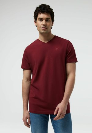 Mężczyzna w gładkiej bordowej koszulce z krótkim rękawem i dekoltem w serek oraz niebieskich dżinsach stoi na jasnym, jednolitym tle.