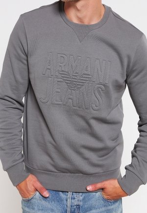Mężczyzna w szarej bluzie Armani Jeans z wytłoczonym logo, w połączeniu z jasnoniebieskimi jeansami, ręce w kieszeniach.