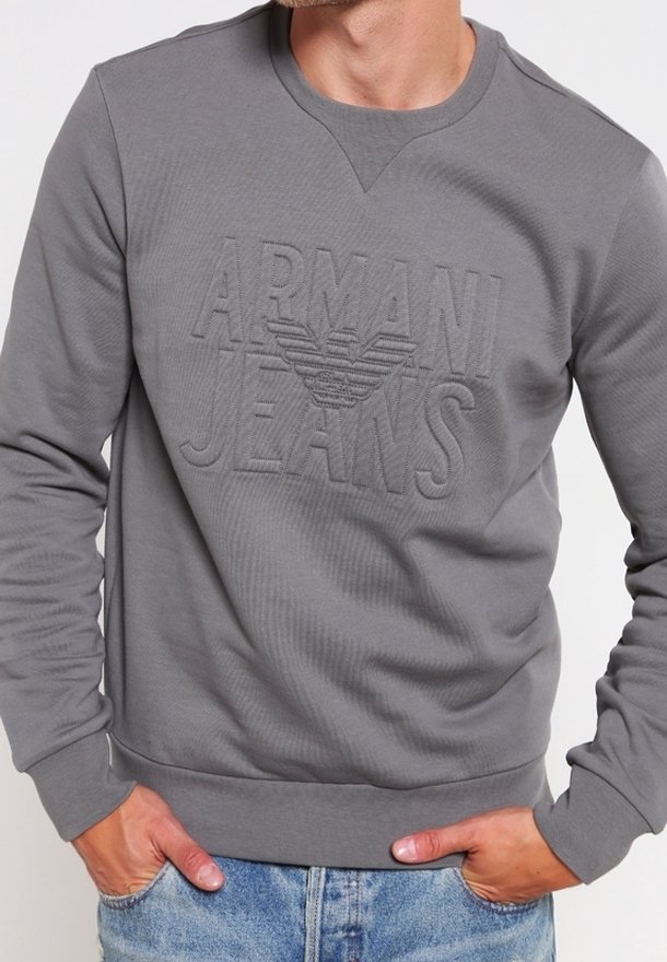Homme portant un sweat-shirt gris Armani Jeans avec logo embossé, associé à un jean denim bleu clair, mains dans les poches.