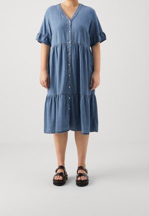 Femme portant une robe en denim bleue boutonnée à l'avant, longueur genou, avec des manches courtes à volants et des sandales noires à bouts ouverts, debout sur un fond uni.