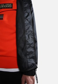 Zwarte nylonjack met meshpaneel, met een oranje sweatshirt eronder. Inclusief ritsen en elastische manchetten voor een verstelbare pasvorm.