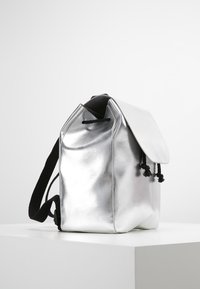 Sac à dos en métal argenté avec une finition lisse, fermeture à cordon de serrage, et bretelles noires. Le design comprend un rabat et une forme structurée.