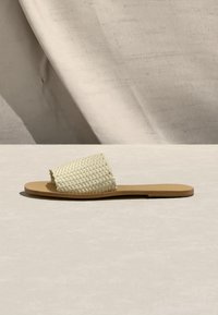 Φλατ καφέ σανδάλι με υφασμάτινο κρεμ λουράκι, εμφανιζόμενο σε φωτεινή επιφάνεια με απαλό ύφασμα φόντο.