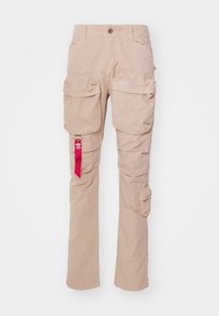Beżowe spodnie cargo z dużymi przednimi kieszeniami, małą kieszonką boczną, zapięciem na guzik oraz czerwoną metką wiszącą z lewej kieszeni.