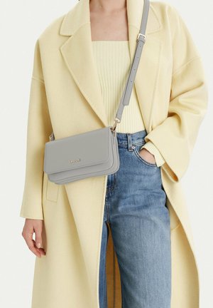 Persona vistiendo un abrigo amarillo pálido, top acanalado, jeans azules y un bolso bandolera gris claro con el logo dorado "LASOCKI".