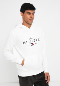 Hvit bomull hettegenser med lommer foran, snøring i hetten og trykt logo i svart og rødt over brystet. Strukturert stoff.