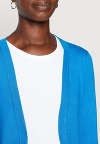 Blå cardigan med en mjuk textur, öppen frontdesign och långa ärmar, lagrad över en vit t-shirt med rund halsringning.