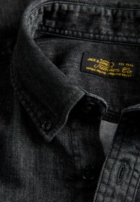 Primo piano del colletto di una camicia di jeans nera con bottoni neri e un'etichetta gialla "Jack & Jones Premium Co." all'interno.