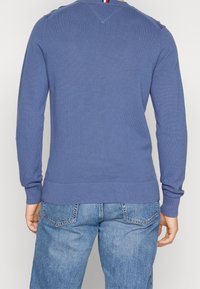 Blå stickad tröja med långa ärmar, strukturerat mönster, ribbade mudd och fåll, samt V-formad detalj på baksidan. Bärs med ljusblå jeans.