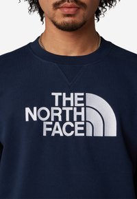Felpa blu navy con colletto tondo a coste, caratterizzata da un logo ricamato bianco prominente "The North Face" sulla parte anteriore.