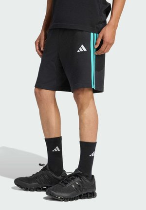 Mężczyzna noszący czarne spodenki Adidas z turkusowymi paskami, czarne skarpetki Adidas oraz czarne buty sportowe, stojący na jasnym, jednolitym tle.