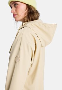 Beige wasserdichte Jacke mit Kapuze, verstellbarem Kordelzug und Roxy-Logo-Patch am Ärmel. Die Textur wirkt glatt und matt.